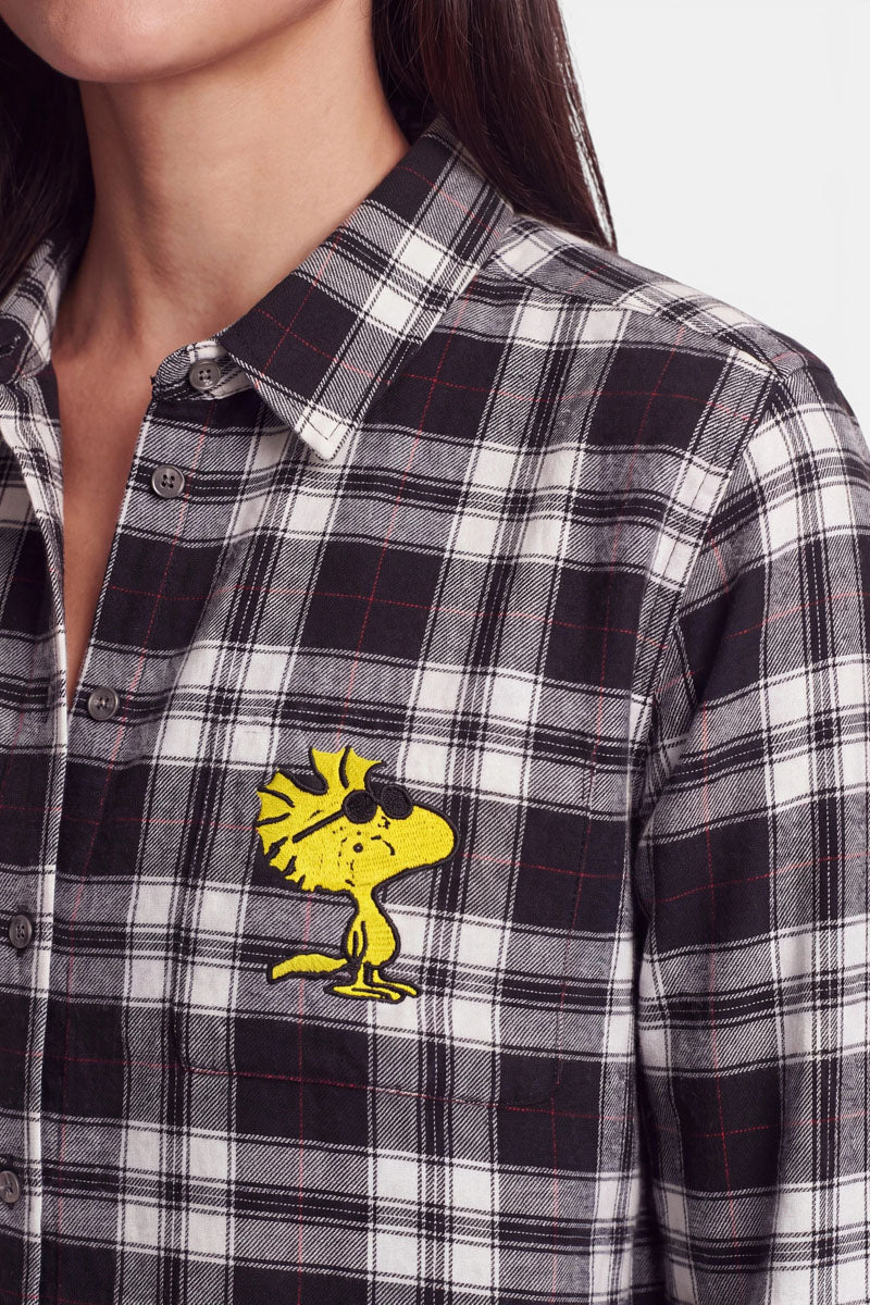 Eine Person trägt eine schwarz-weiß-grau karierte Hemdbluse mit klassischem Karomuster. Auf der linken Brust ist ein gelber Comic-Vogel aufgestickt – Woodstock aus „Peanuts“. Die Bluse ist zugeknöpft, und nur die untere Hälfte des Gesichts und des Oberkörpers der Person sind zu sehen.