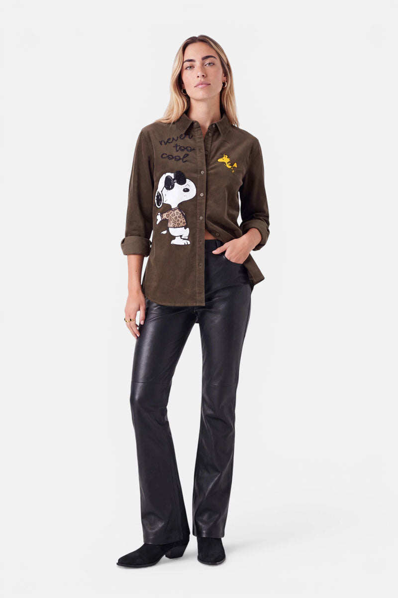 Eine Frau steht mit dem Blick nach vorne und trägt eine braune Hemdbluse aus Cord mit Snoopy-Aufdruck sowie Abbildungen von Snoopy und Woodstock, eine schwarze Lederhose und schwarze Schuhe. Die Ärmel ihrer Bluse sind hochgekrempelt, und ihre linke Hand steckt in der Tasche. Sie hat langes, offenes Haar und einen neutralen Gesichtsausdruck.