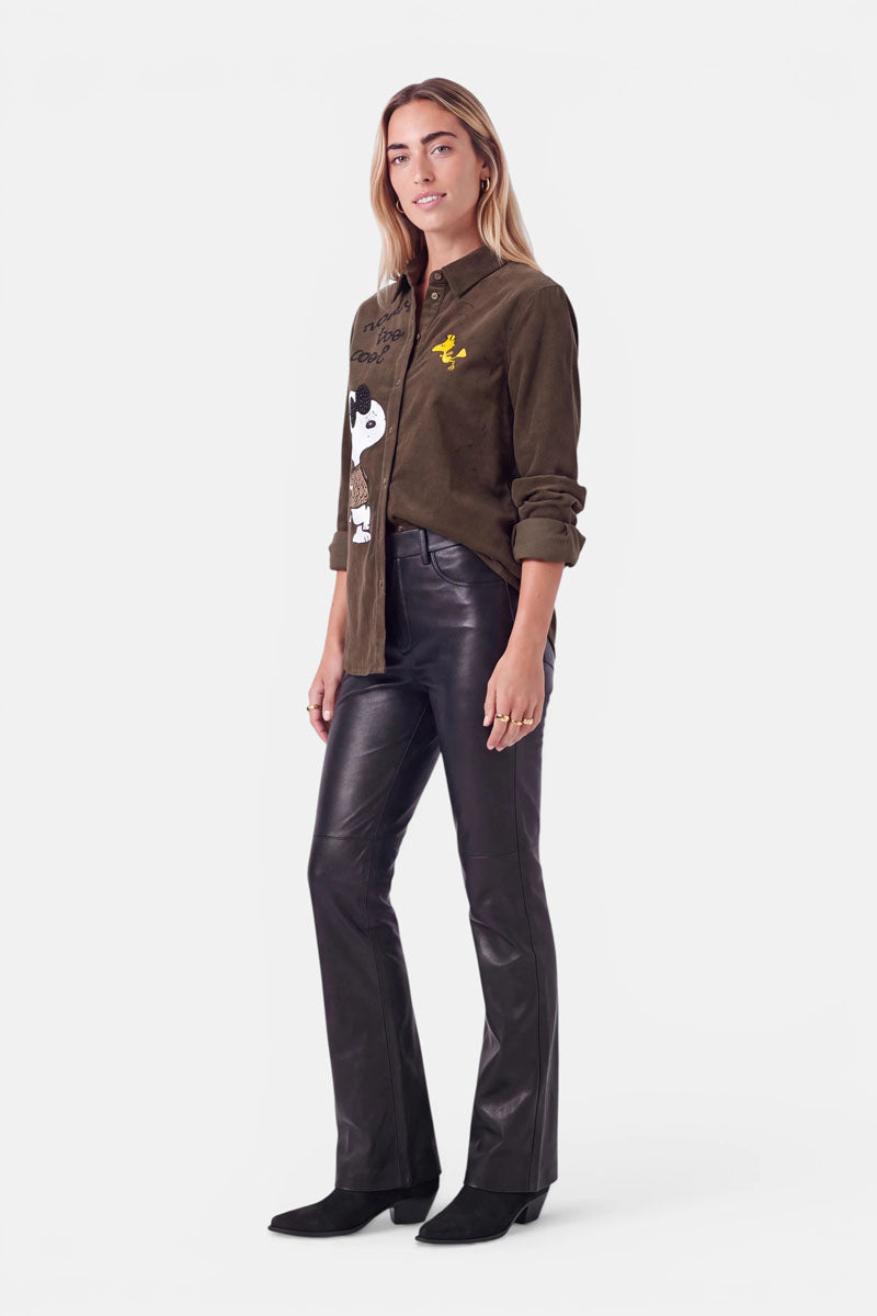 Eine Frau mit langen blonden Haaren trägt eine braune Cord-Hemdbluse mit verspielten Snoopy-Motiven und dezenten Strass-Akzenten, dazu schwarze Lederhosen und schwarze Schuhe. Sie steht in einer entspannten Haltung vor einem schlichten weißen Hintergrund.