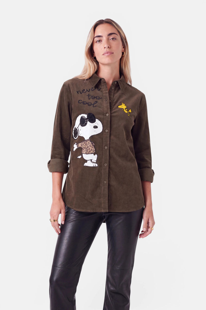 Eine Frau mit langen blonden Haaren trägt eine stilvolle, olivgrüne Cord-Hemdbluse mit einem verspielten Snoopy-Aufdruck, auf dem Woodstock und der Schriftzug „never too cool“ zu sehen sind, elegant kombiniert mit einer schwarzen Lederhose. Sie steht vor einem schlichten weißen Hintergrund, blickt selbstbewusst in die Kamera und hält die Hände an den Seiten.