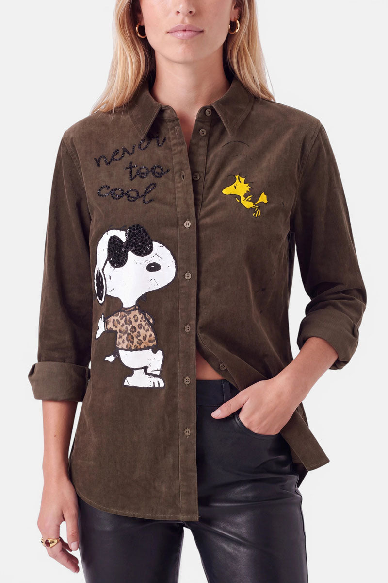 Eine Frau trägt eine olivgrüne Cord-Hemdbluse mit Snoopy-Aufdruck und Woodstock-Aufnähern auf der Vorderseite, dem Schriftzug „never too cool“, Strass-Details und hochgekrempelten Ärmeln. Dazu trägt sie eine schwarze Lederhose und hat langes blondes Haar, goldene Ohrringe und Ringe.
