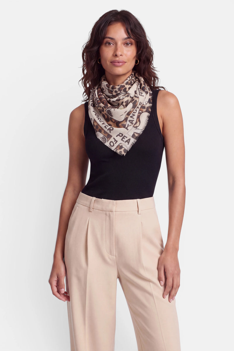 Eine Frau mit welligem braunem Haar trägt ein ärmelloses schwarzes Oberteil, eine beige Faltenhose und einen stilvollen Peanuts-Schal aus einer Baumwoll-Woll-Mischung mit schwarzer Aufschrift. Sie steht mit dem Blick nach vorne vor einem schlichten weißen Hintergrund, einen Arm entspannt an der Seite.