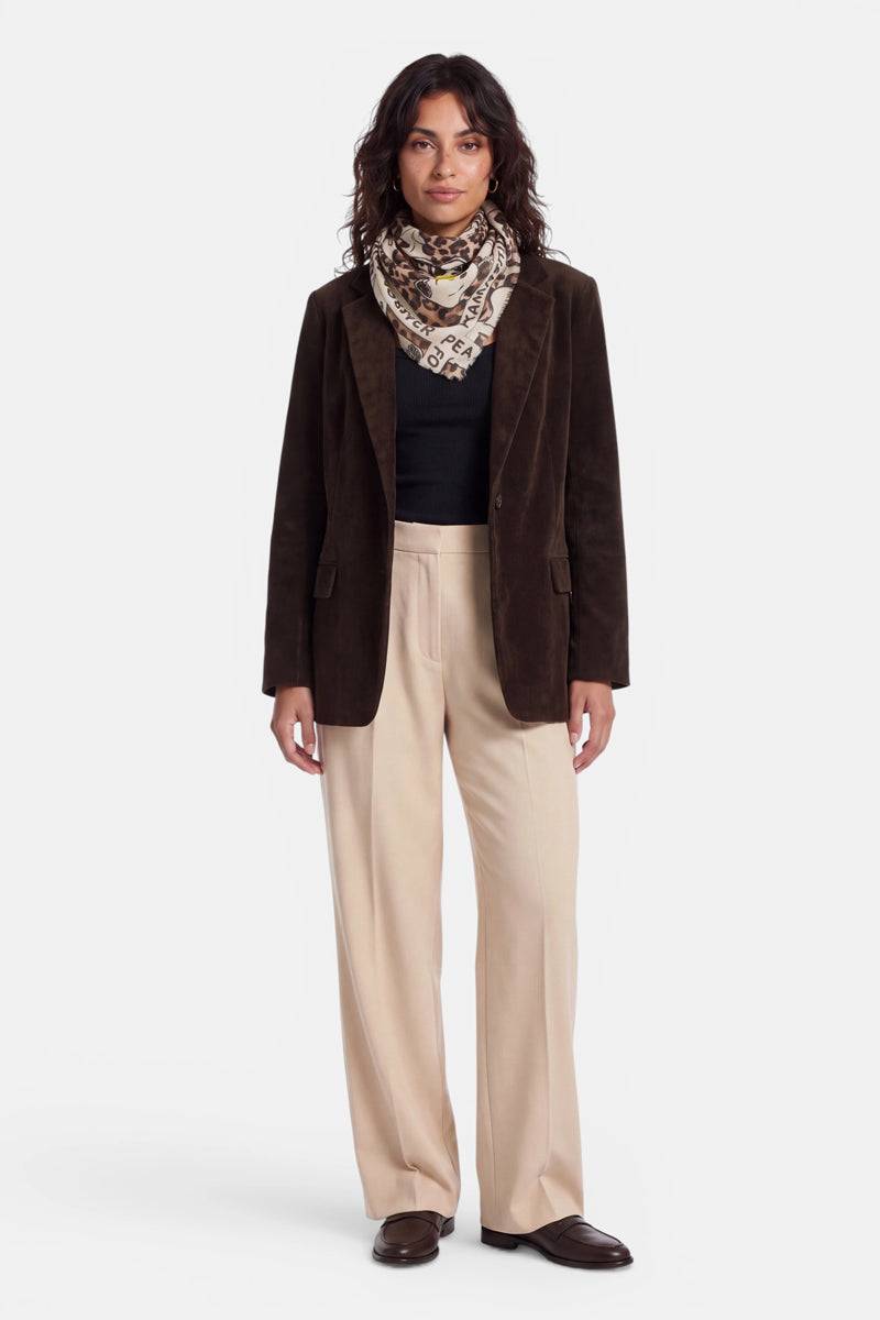 Eine Frau steht mit dem Blick nach vorne gerichtet da. Sie trägt einen dunkelbraunen Blazer, ein schwarzes Oberteil, eine beige Hose mit weitem Bein aus einem Baumwoll-Woll-Mix, braune Schuhe und einen „Peanuts“-Schal, der um ihren Hals drapiert ist. Sie hat welliges dunkles Haar und einen neutralen Gesichtsausdruck, vor einem schlichten weißen Hintergrund.