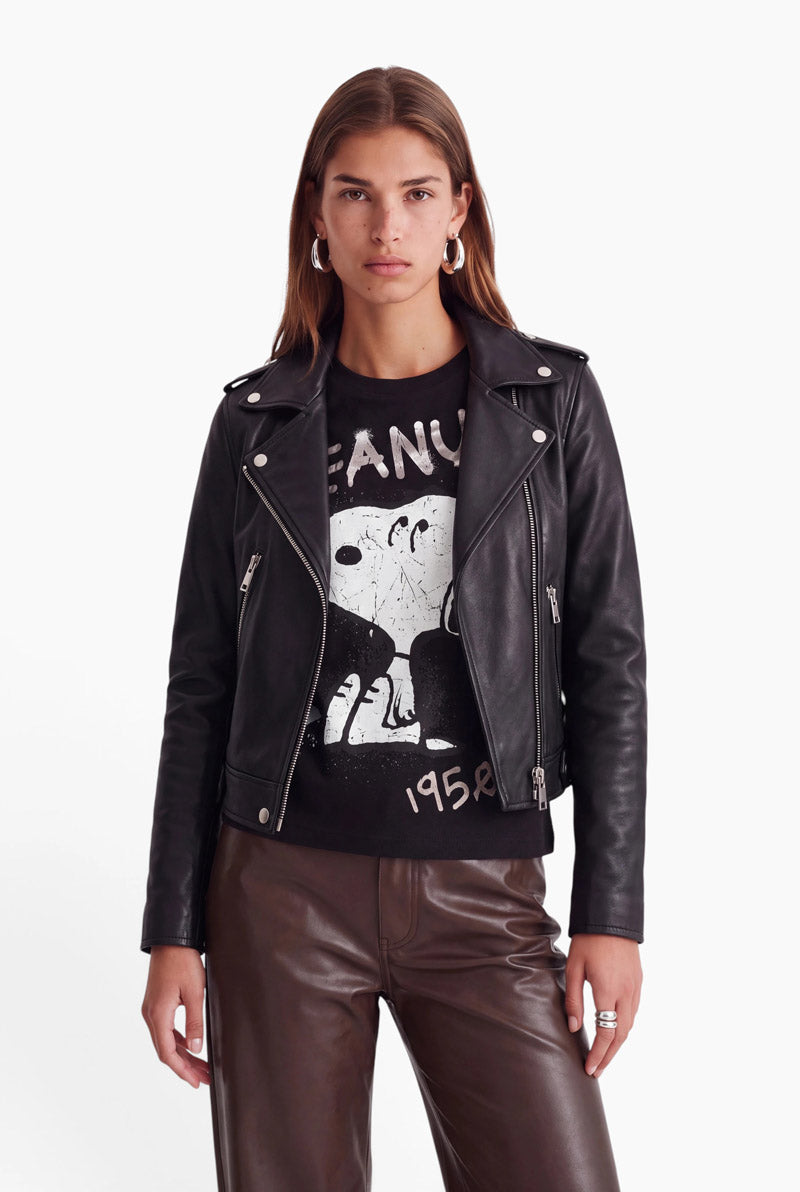 Eine Frau mit langen, hellbraunen Haaren trägt eine schwarze Leder-Motorradjacke über einem Peanuts 1950-T-Shirt aus Baumwolle. Zu diesem Outfit kombiniert sie eine braune Hose und silberne Creolen vor einem schlichten weißen Hintergrund.