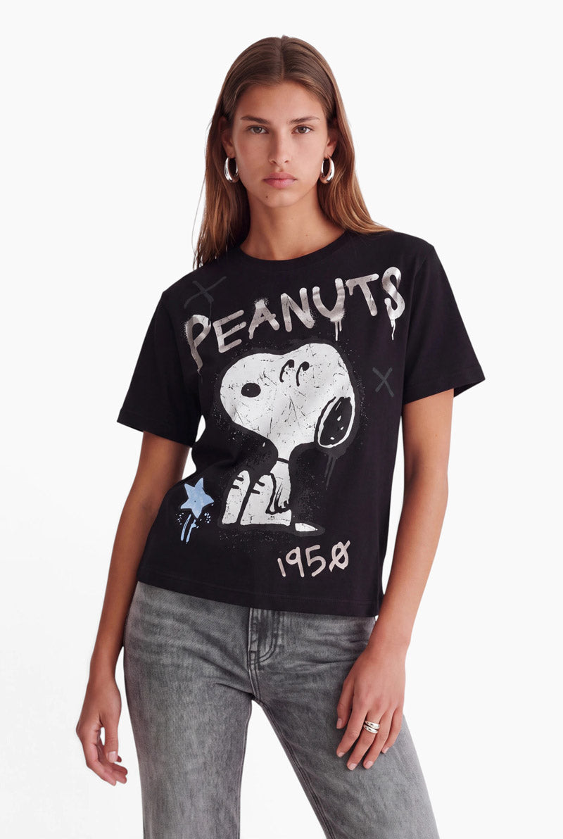 Eine Frau mit langen braunen Haaren und silbernen Creolen trägt ein schwarzes Baumwoll-T-Shirt mit Snoopy-Aufdruck, auf dem in weißer Schrift PEANUTS und 1950 zu lesen sind. Sie trägt eine graue Jeans und steht vor einem schlichten weißen Hintergrund.