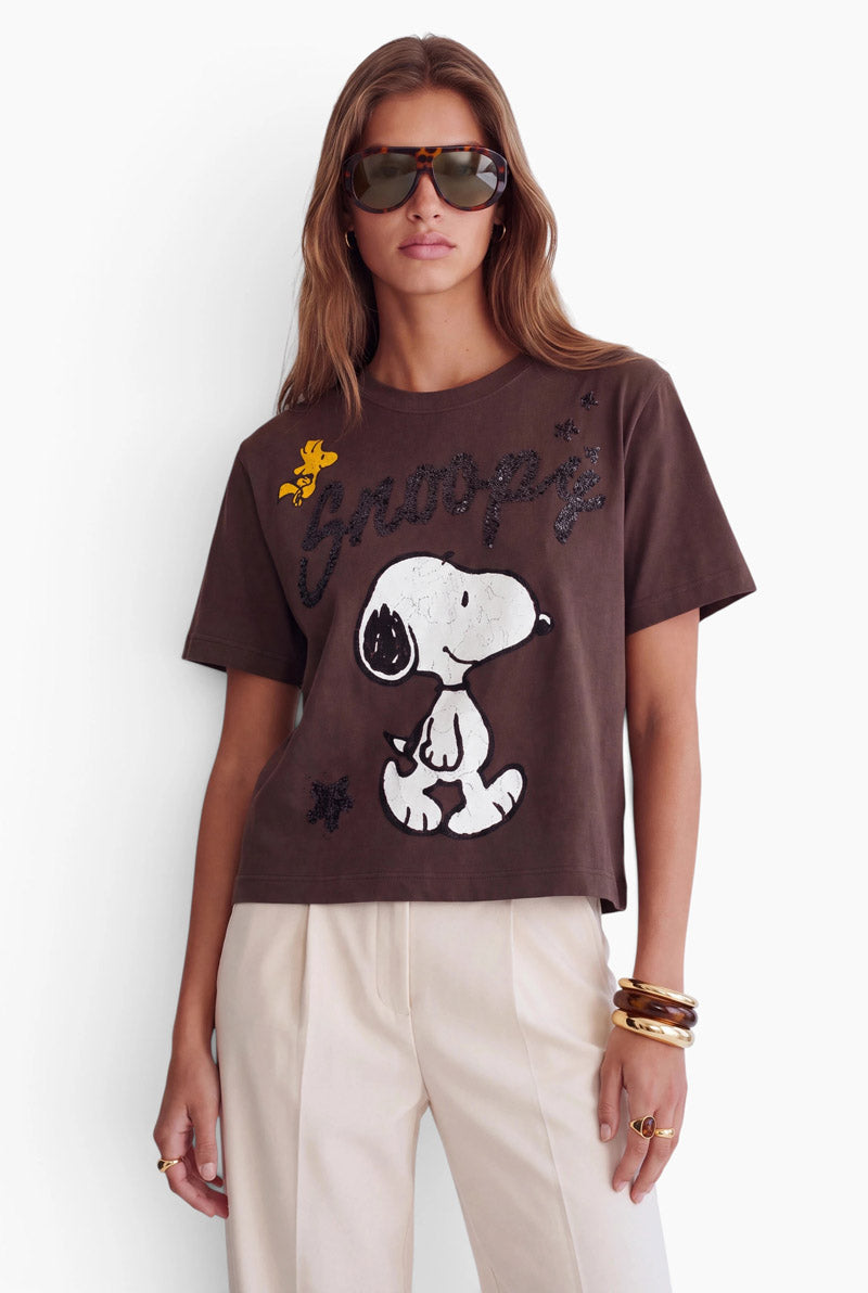 Eine Frau trägt ein braunes Snoopy-T-Shirt aus Baumwolle, auf dem eine große Grafik der Figur und ihr Name in Schreibschrift abgebildet sind. Sie trägt eine übergroße Sonnenbrille in Schildpattoptik, eine cremefarbene Hose sowie mehrere goldene und dunkelbraune Armbänder übereinander.