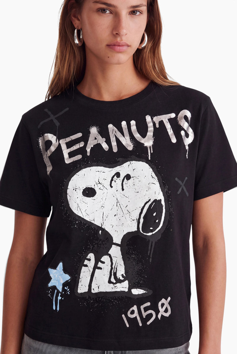 Eine Frau trägt ein schwarzes Baumwoll-T-Shirt mit einem Snoopy-Aufdruck im Graffiti-Stil, der wie abgenutzt wirkt. Das Motiv zeigt das Wort PEANUTS, die Jahreszahl 1958, einen Stern und ein X, alles in einem verwitterten weißen Farbeffekt auf dem dunklen Stoff.