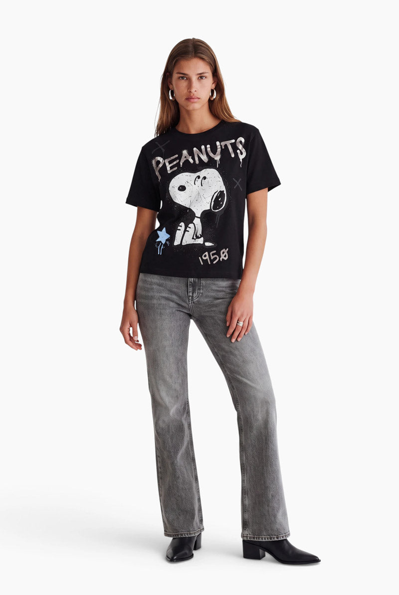 Eine Frau posiert in einem schwarzen Snoopy-T-Shirt aus Baumwolle mit der Aufschrift PEANUTS 1958. Sie trägt eine graue Jeans mit geradem Bein und schwarze Stiefel mit spitzer Zehenpartie und steht vor einem schlichten weißen Hintergrund.