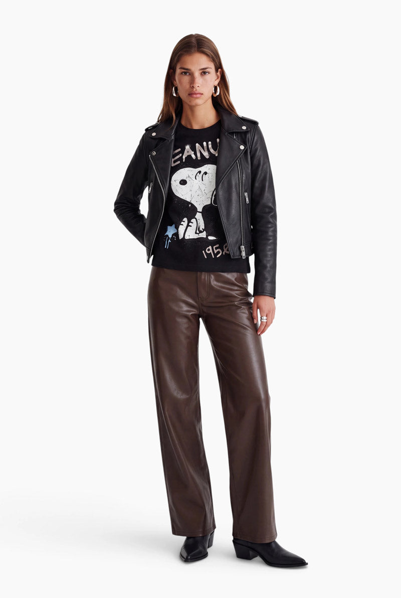Eine Frau trägt eine schwarze Leder-Motorradjacke über einem T-Shirt mit einem Snoopy-Motiv aus den 1950er Jahren. Dazu kombiniert sie eine dunkelbraune Hose mit weitem Bein und schwarze Lederstiefel.