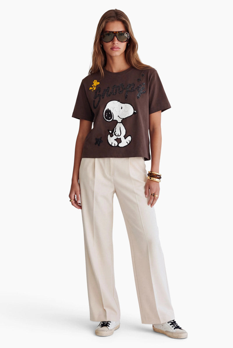 Ein Model trägt ein braunes Baumwoll-T-Shirt mit Snoopy-Motiv im Crop-Schnitt und dazu eine weiße Hose mit weitem Bein. Vor einem schlichten weißen Hintergrund trägt sie eine Sonnenbrille in Schildpattoptik, mehrere goldene Armbänder übereinander und weiße Sneaker.