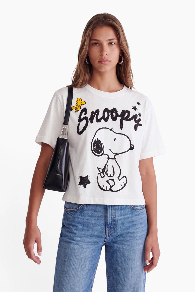 Eine Frau mit langen braunen Haaren trägt ein weißes Snoopy-T-Shirt mit einer schwarz-weißen Illustration, dem Schriftzug Snoopy in Schreibschrift und einem gelben Woodstock. Sie trägt eine schwarze Umhängetasche und blaue Jeans.