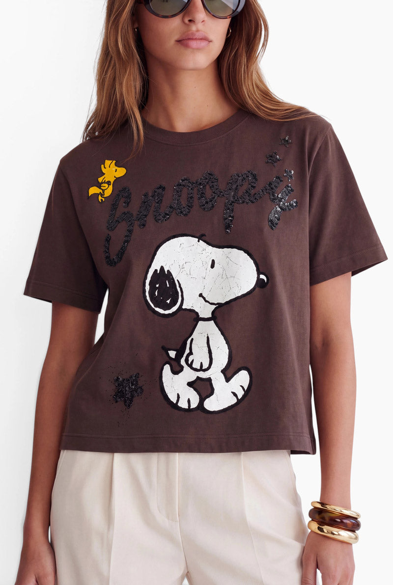 Eine Frau trägt ein braunes Snoopy-T-Shirt aus Baumwolle mit einem großen Snoopy-Motiv und dem Schriftzug Snoopy aus schwarzen Pailletten. Das Shirt ist außerdem mit einer kleinen gelben Woodstock-Figur, schwarzen Pailletten-Sternen sowie klobigen Armbändern in Gold und Schildpatt verziert.