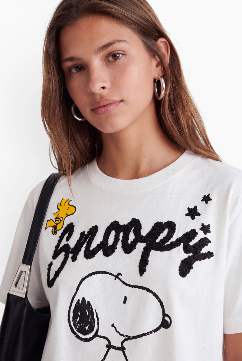 Eine Frau mit langen braunen Haaren trägt ein weißes Snoopy-T-Shirt aus Baumwolle mit Rundhalsausschnitt, das mit einem schwarzen Motiv, strukturiertem Schriftzug und einem gelben Woodstock verziert ist. Sie trägt silberne Creolen und eine schwarze Umhängetasche.