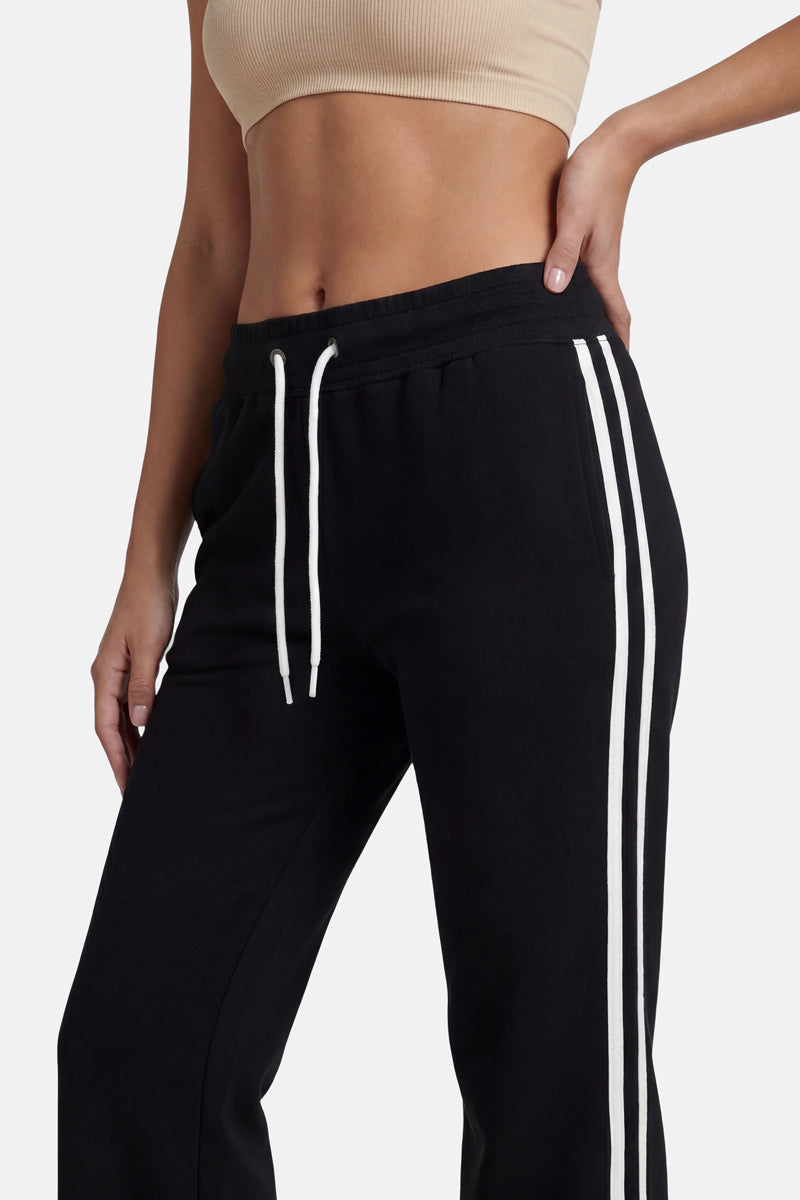 Eine Frau trägt eine schwarze Baumwoll-Jogginghose aus der „Princess Goes Hollywood“-Limited-Edition mit zwei weißen Seitenstreifen und einem elastischen Bund mit weißen Kordeln, dazu ein beiges, geripptes Crop-Top. Sie steht vor einem einfarbigen hellgrauen Hintergrund.