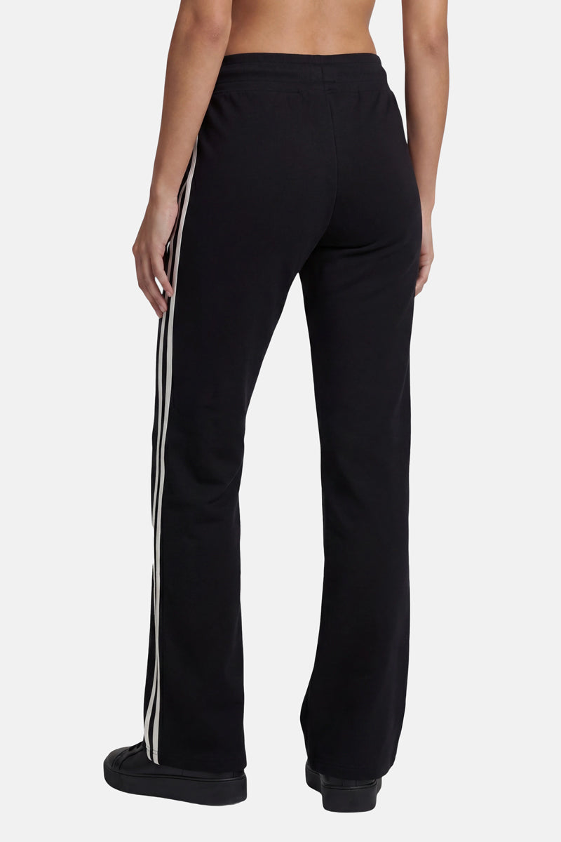 Eine Person steht mit dem Rücken zur Kamera und trägt eine schwarze Sporthose in geradem Schnitt aus der limitierten „Princess Goes Hollywood“-Kollektion mit zwei weißen Seitenstreifen. Diese aus hochwertiger Baumwolle gefertigte Jogginghose sitzt in der Taille und wird mit schwarzen Schuhen kombiniert.