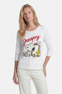 Eine Frau mit langen blonden Haaren trägt ein „Princess Goes Hollywood Limited Edition Snoopy“-Shirt – ein weißes Baumwolloberteil mit Dreiviertelärmeln, auf dem Snoopy und Woodstock an einem Limonadenstand abgebildet sind. Dazu trägt sie eine weiße Hose vor einem schlichten weißen Hintergrund.