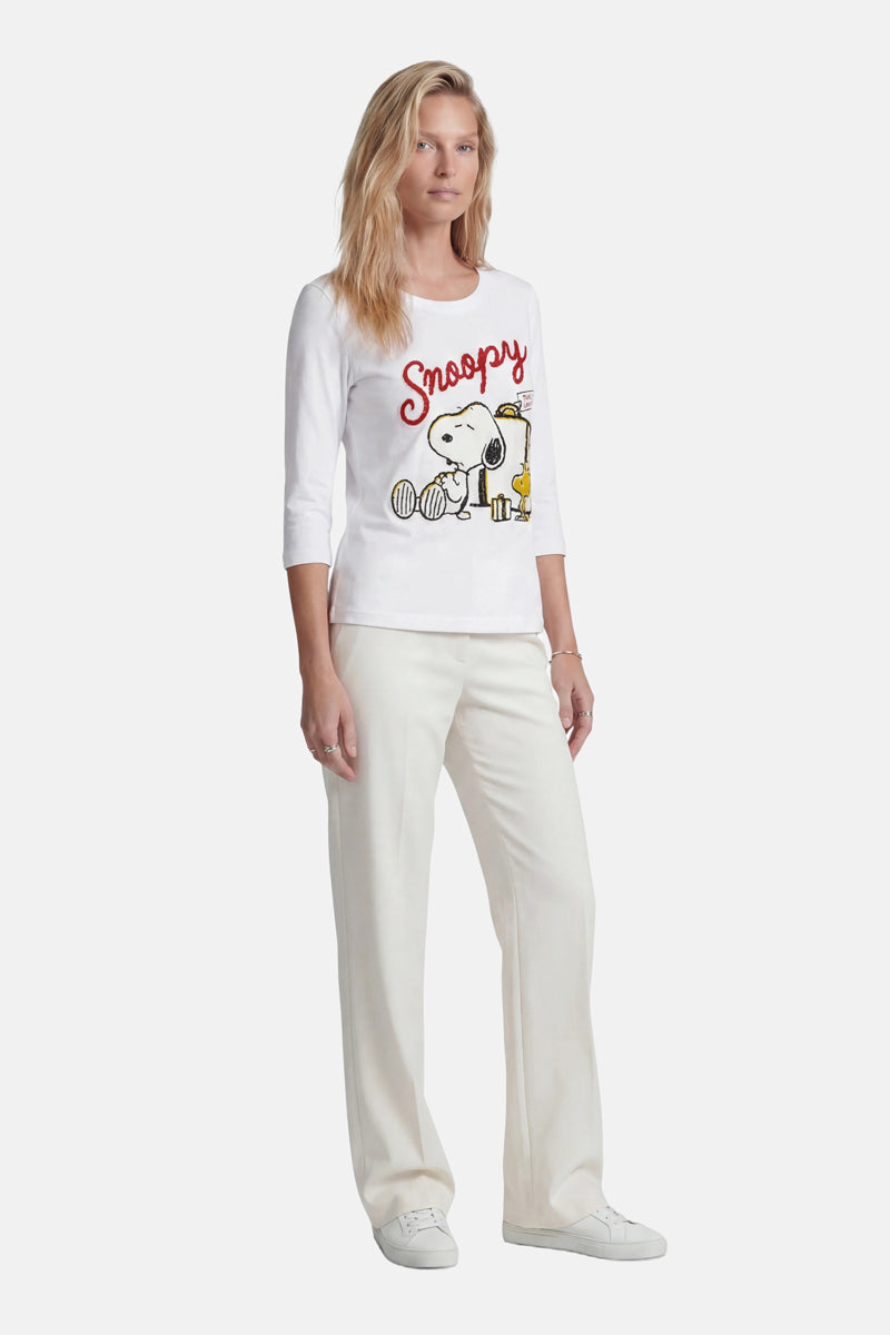 Eine Frau mit langen blonden Haaren trägt ein „Princess Goes Hollywood“-Snoopy-Shirt aus Baumwolle in limitierter Auflage, dazu weiße Hosen und Turnschuhe. Sie steht mit dem Gesicht zur Kamera vor einem schlichten, hellen Hintergrund.