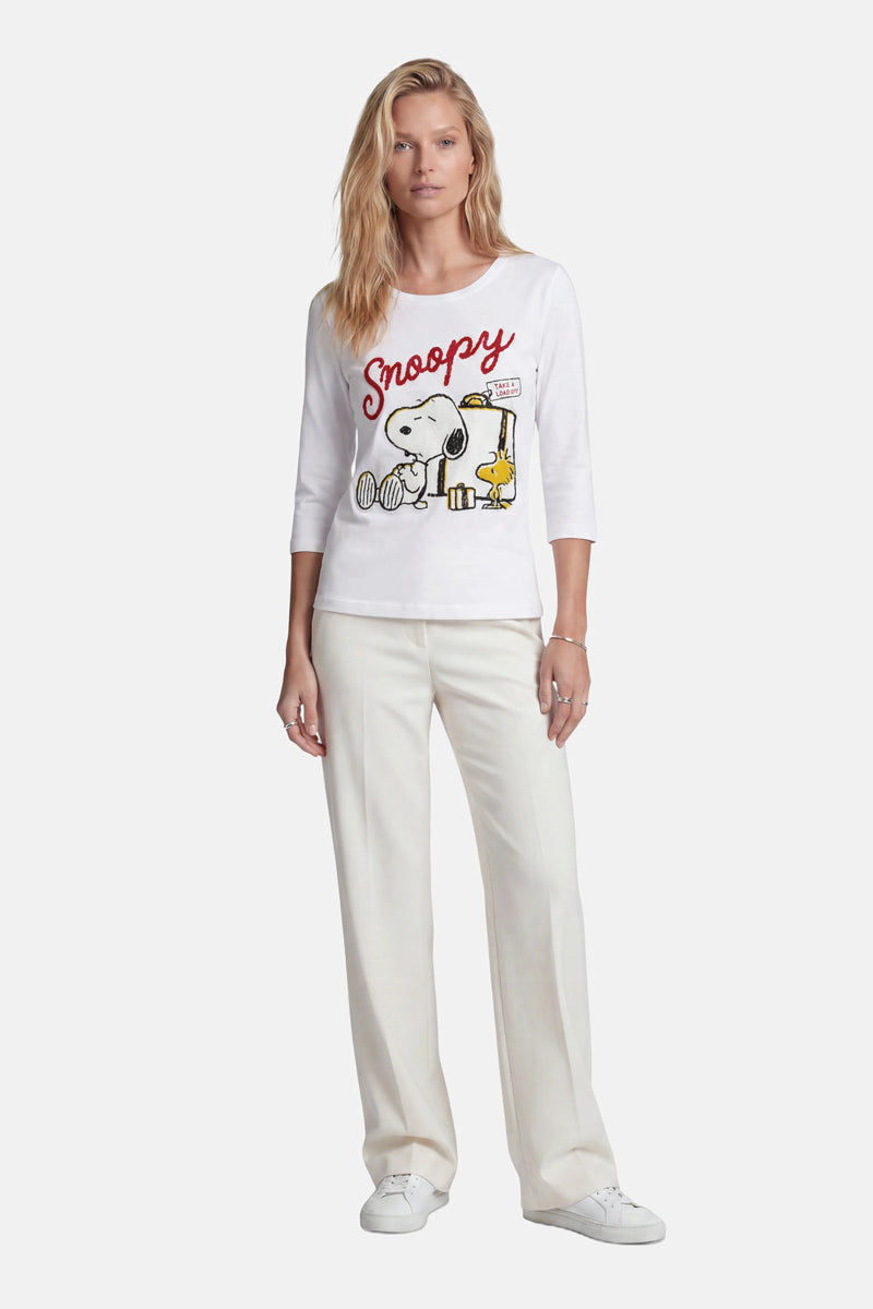 Eine blonde Frau trägt ein Snoopy-Shirt aus der „Princess Goes Hollywood“-Limited-Edition aus Baumwolle, auf dem Snoopy und Woodstock abgebildet sind und der Schriftzug „Snoopy“ in Rot zu sehen ist. Dazu trägt sie eine cremefarbene Hose mit weitem Bein und weiße Sneaker vor einem hellen Hintergrund.