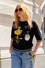 Eine Frau mit langen, welligen blonden Haaren trägt eine große braune Sonnenbrille und einen schwarzen Strickpullover „Woodstock-Intarsia“ in limitierter Auflage von Princess Goes Hollywood, auf dem ein gelber Comic-Vogel und der Schriftzug „TRAVEL DAY“ zu sehen sind. Sie steht vor einem Steingebäude, die Hände in den Taschen.