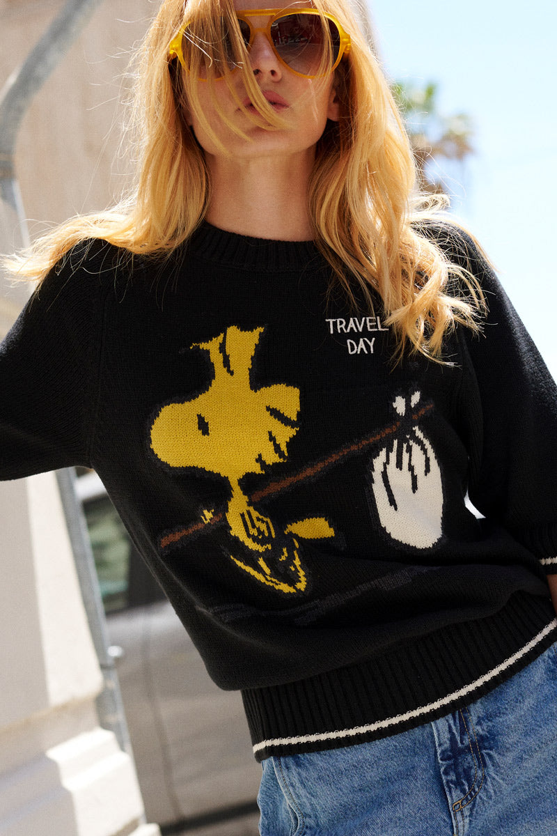 Eine Frau mit langen blonden Haaren trägt eine orangefarbene Sonnenbrille und einen schwarzen Strickpullover der Limited Edition „Princess Goes Hollywood“ mit gelber Woodstock-Intarsia und hält einen weißen „TRAVEL DAY“-Beutel in der Hand. Sie steht im Freien, hinter ihr sind ein Auto und Gebäude verschwommen zu sehen.