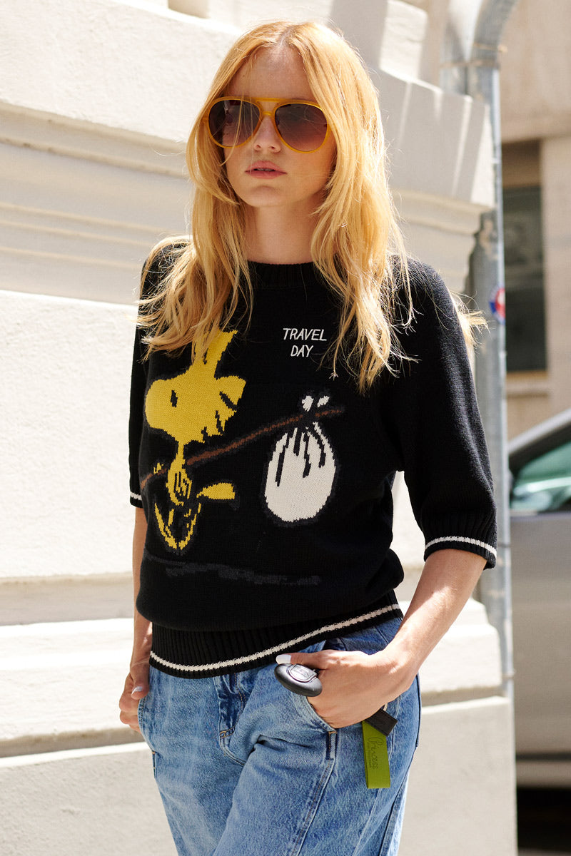 Eine Frau mit langen blonden Haaren trägt eine große Pilotenbrille und einen schwarzen Strickpullover aus der „Princess Goes Hollywood“-Limited-Edition mit Woodstock-Intarsien und der Aufschrift „TRAVEL DAY“, dazu hellblaue Jeans; sie steht draußen im Sonnenlicht neben einem weißen Gebäude.