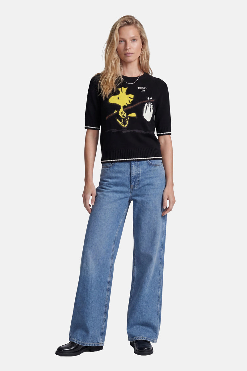 Eine Frau mit langen blonden Haaren trägt einen schwarzen Strickpullover der Limited Edition „Princess Goes Hollywood“ mit gelbem Woodstock-Intarsia-Muster und Schriftzug, eine lockere blaue Jeans mit weitem Bein und schwarze Schuhe. Sie steht mit dem Blick nach vorne vor einem schlichten weißen Hintergrund.