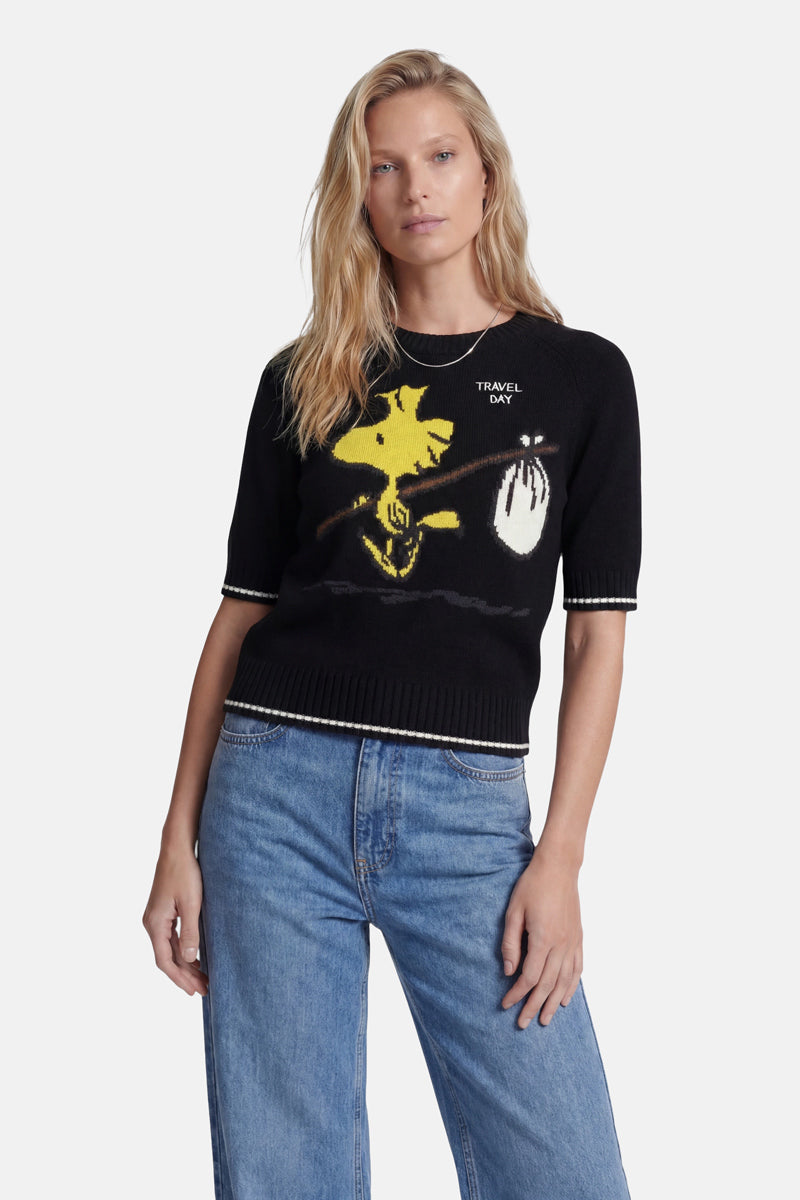 Eine Frau mit langen blonden Haaren trägt einen schwarzen Strickpullover von „Princess Goes Hollywood“ im Woodstock-Intarsia-Stil mit der „Peanuts“-Figur und dem Schriftzug „TRAVEL DAY“, dazu eine hellblaue Jeans mit hoher Taille vor einem schlichten weißen Hintergrund.