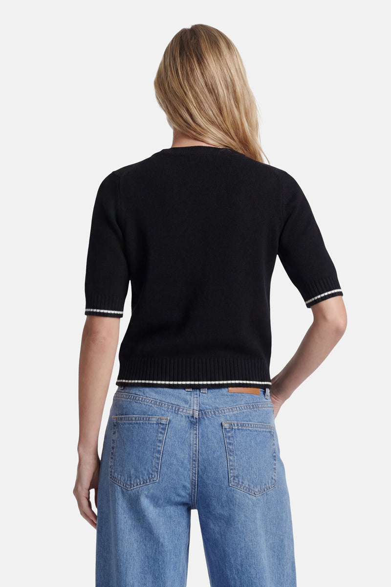 Eine Person mit langen, hellbraunen Haaren, von hinten gesehen, trägt einen schwarzen „Princess Goes Hollywood“-Pullover im Woodstock-Intarsia-Stil mit weißen Bordüren an den Ärmeln und am Saum, dazu eine blaue Jeans mit hoher Taille vor einem hellgrauen Hintergrund.