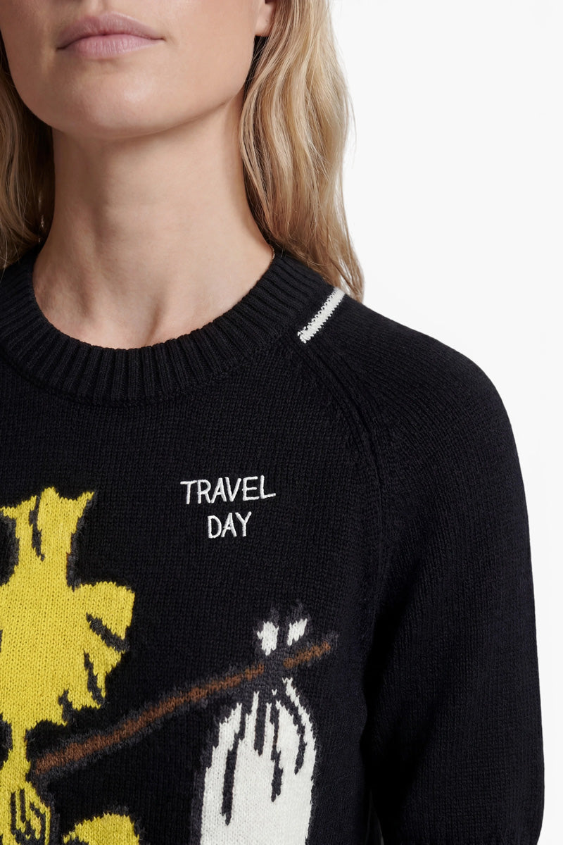 Eine hellhäutige Frau mit welligem blondem Haar trägt einen schwarzen Strickpullover aus der „Princess Goes Hollywood“-Limited-Edition mit der Aufschrift „TRAVEL DAY“ in Weiß und einer gelb-weißen Cartoon-Grafik. Das Bild zeigt sie von den Schultern aufwärts vor einem schlichten weißen Hintergrund.