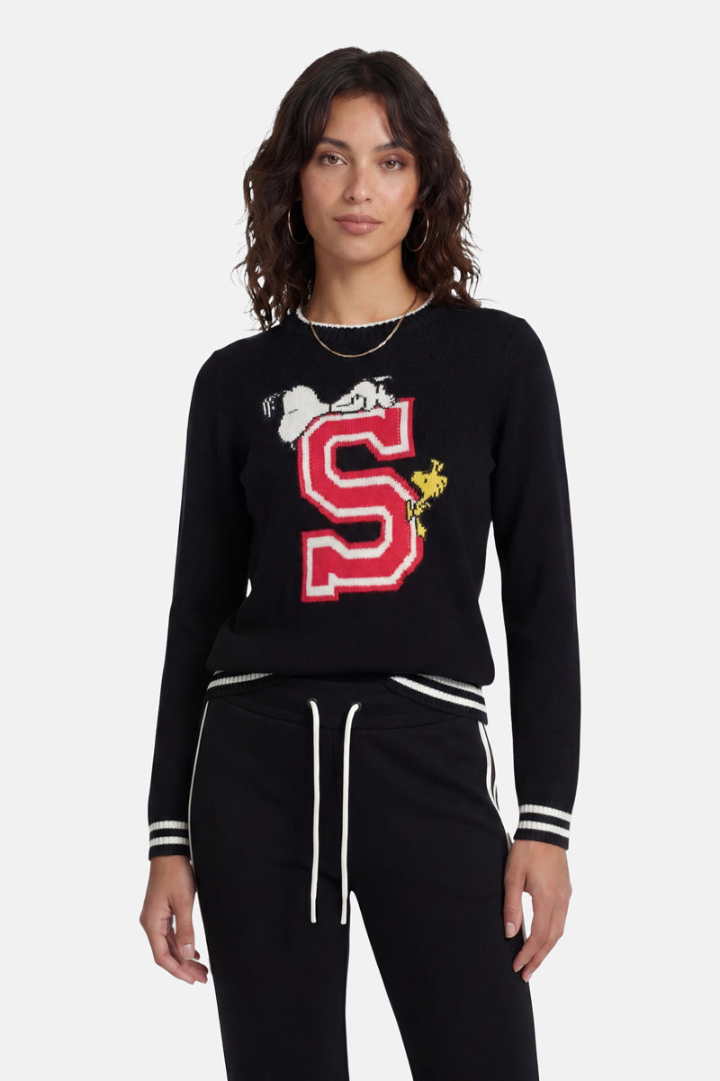 Eine Frau mit welligem braunem Haar trägt einen schwarzen Strickpullover aus der „Princess Goes Hollywood“-Limited-Edition mit einem großen roten „S“, darauf liegend Snoopy als Intarsienmotiv und daneben Woodstock, dazu eine schwarze Hose mit weiß gestreiften Bündchen und eine Kettenhalskette.