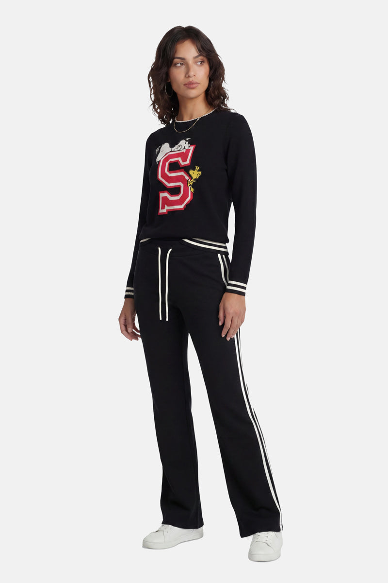 Eine Frau trägt ein schwarzes Langarm-Oberteil aus der „Princess Goes Hollywood“-Limited-Edition mit einem großen roten „S“ und einem Snoopy-&amp;-Woodstock-Motiv, dazu eine passende schwarze Hose mit weißen Seitenstreifen und Kordelzug. Abgerundet wird der Look durch weiße Sneaker vor einem schlichten weißen Hintergrund.