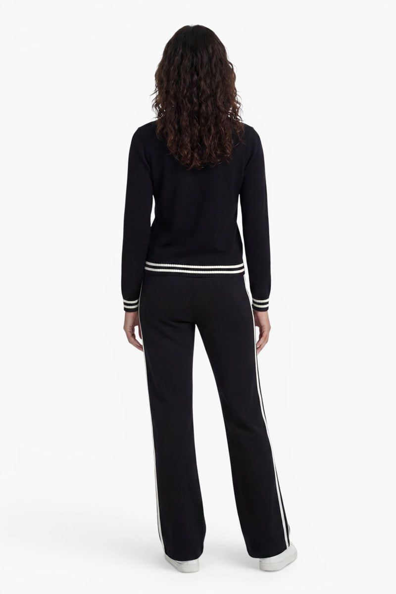 Eine Person mit mittellangem, lockigem Haar ist von hinten zu sehen. Sie trägt ein schwarzes Langarm-Oberteil aus der „Princess Goes Hollywood“-Limited-Edition und eine dazu passende schwarze Hose mit weißen Streifen an den Seiten und an den Bündchen, dazu weiße Sneaker vor einem schlichten weißen Hintergrund.