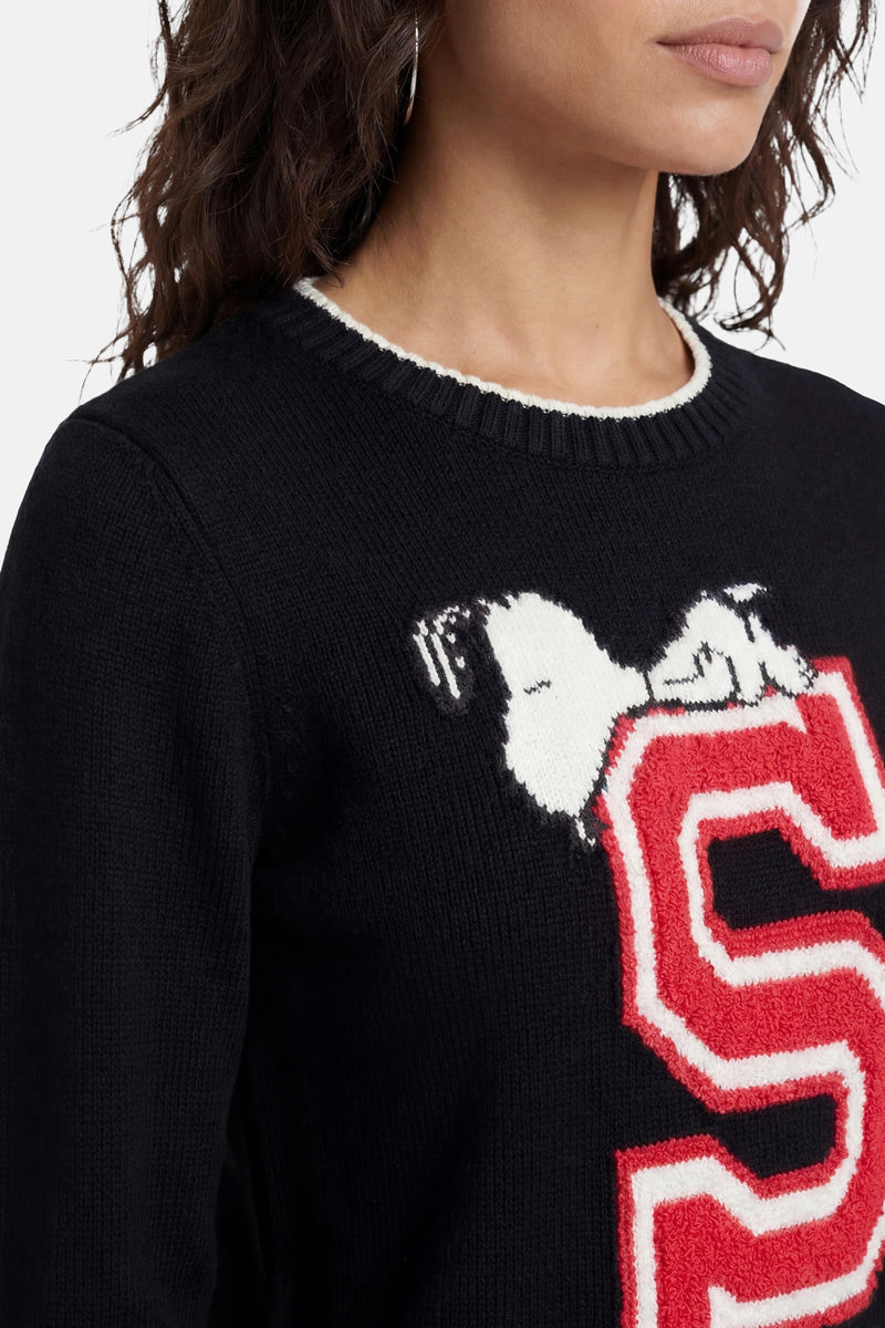 Eine Frau trägt einen schwarzen „Princess Goes Hollywood“-Snoopy-Intarsia-Strickpullover, auf dem ein weiß-roter Buchstabe „S“ zu sehen ist, auf dem Snoopy liegt. Vor einem schlichten weißen Hintergrund sind nur ihr unterer Gesichtsbereich, ihre Haare und ihr Oberkörper zu sehen.