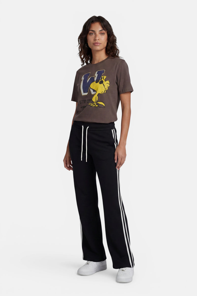 Eine Frau mit welligem, dunklem Haar steht vor einem weißen Hintergrund. Sie trägt ein braunes Baumwoll-T-Shirt der limitierten Auflage „Princess Goes Hollywood“ mit Woodstock-Aufdruck und einem „W“, eine schwarze Jogginghose mit weißen Streifen und Kordelzug sowie weiße Sneaker. Ihr Gesichtsausdruck ist neutral.