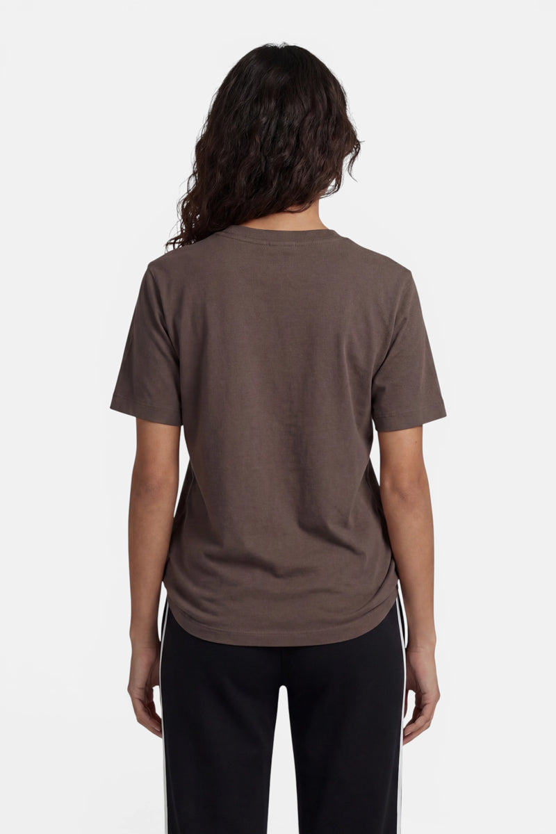 Eine Person mit langen, gewellten Haaren steht mit dem Rücken zur Kamera und trägt ein schlichtes braunes „Princess Goes Hollywood“-Baumwoll-T-Shirt sowie eine schwarze Hose mit weißen Seitenstreifen vor einem schlichten weißen Hintergrund.