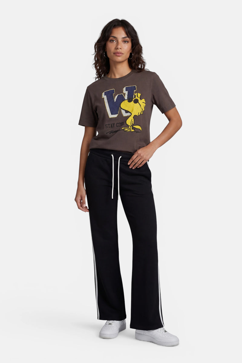 Eine Frau mit gewelltem Haar und einem entspannten Gesichtsausdruck blickt nach vorne. Sie trägt ein T-Shirt der Limited Edition „Princess Goes Hollywood“ in verblasstem Braun mit einem großen blauen „W“ und einem gelben Woodstock-Aufdruck, eine schwarze Jogginghose mit Kordelzug und weißen Seitenstreifen sowie weiße Turnschuhe.
