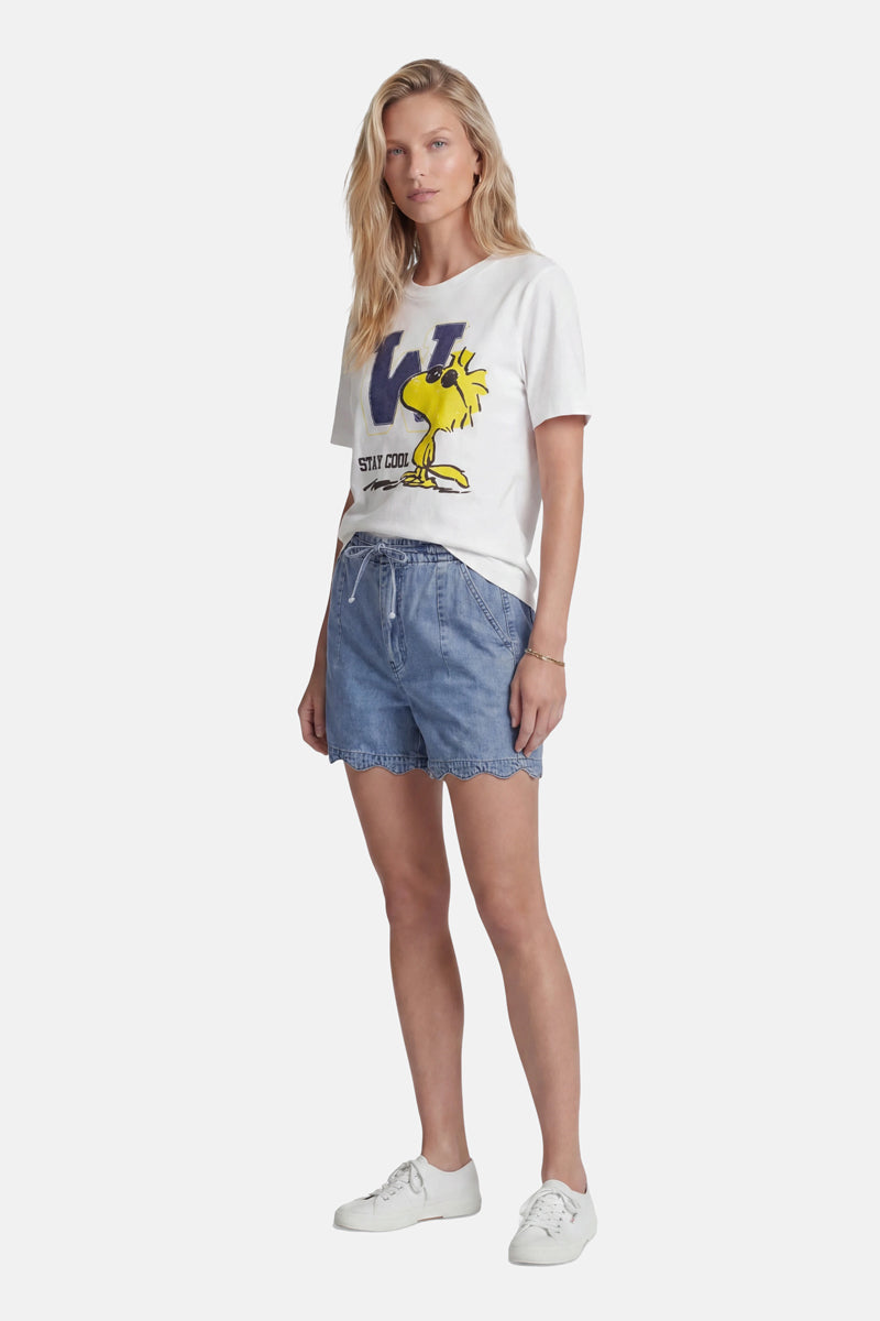 Eine Frau mit langen blonden Haaren trägt ein weißes Baumwoll-T-Shirt der Limited Edition „Princess Goes Hollywood“ mit einem auffälligen gelben Woodstock-Aufdruck und dem Schriftzug „Stay Cool“. Dazu trägt sie blaue Jeans-Shorts mit Kordelzug und weiße Sneaker und steht vor einem einfarbigen Hintergrund.