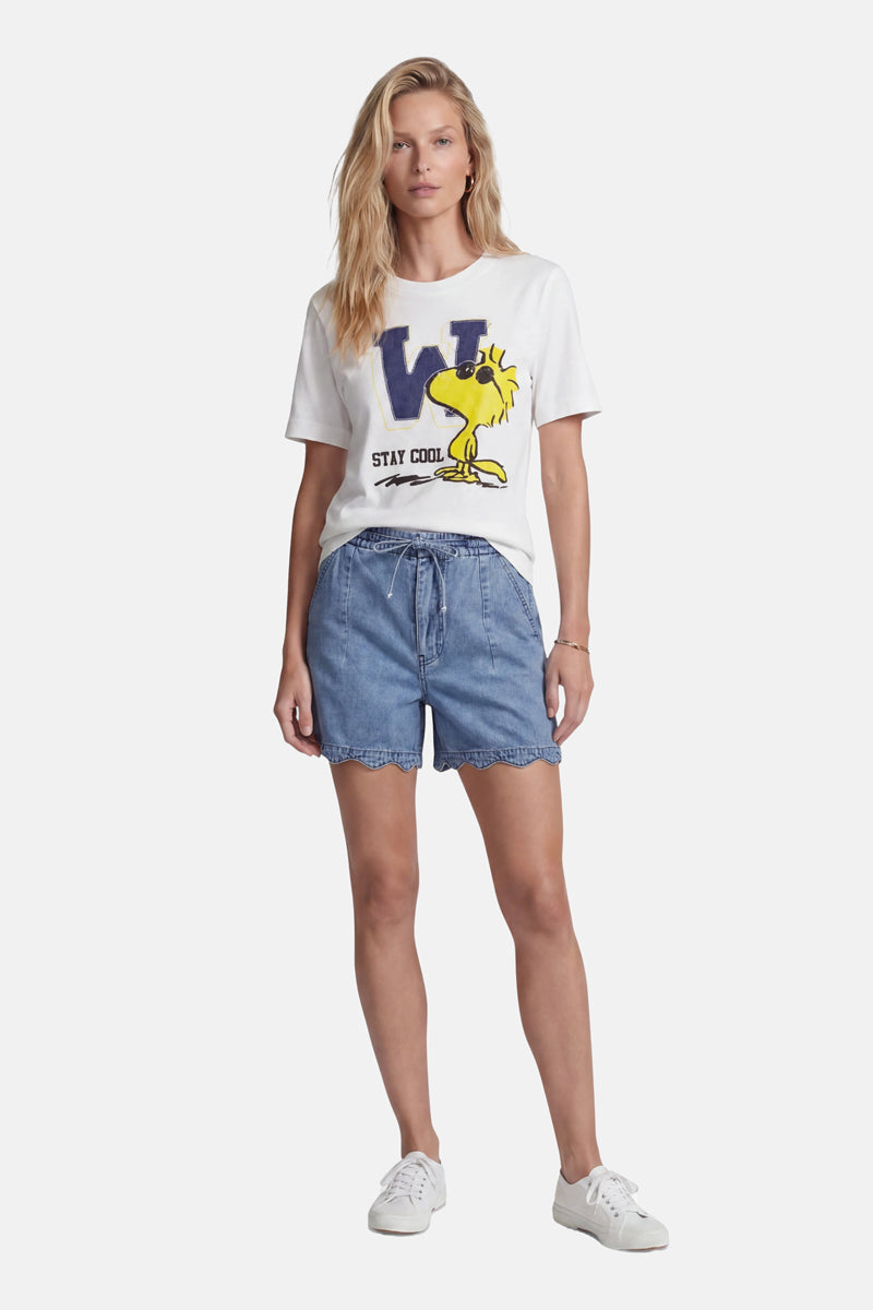 Eine Frau mit welligem blondem Haar und ausdruckslosem Gesicht trägt ein „Princess Goes Hollywood Limited Edition“-T-Shirt aus Baumwolle mit einem großen blauen „W“, einem Woodstock-Aufdruck und dem Schriftzug „STAY COOL“, das sie in Jeansshorts mit Kordelzug und gewellten Säumen gesteckt hat, dazu weiße Sneaker.