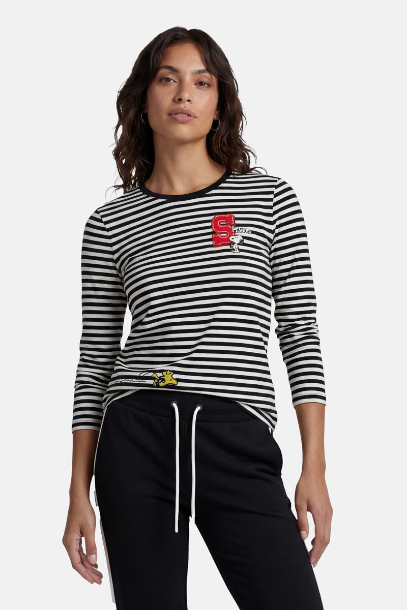 Eine Frau trägt ein gestreiftes Langarmshirt aus der „Princess Goes Hollywood“-Limited-Edition mit einem rot-weißen Aufnäher und einer dezenten Snoopy-Stickerei auf der Brust, dazu eine schwarze Hose mit Kordelzug. Ihr welliges dunkles Haar hebt sich deutlich vom hellen Hintergrund ab.