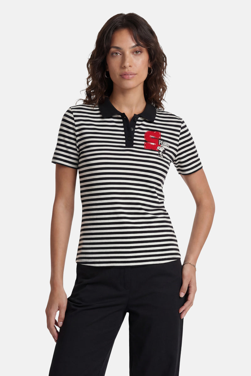 Eine Frau mit welligem braunem Haar trägt ein gestreiftes T-Shirt aus der „Princess Goes Hollywood“-Limited-Edition mit schwarzem Kragen, einem roten „S“-Aufnäher und einer Snoopy-Stickerei auf der linken Brustseite. Dazu trägt sie eine schwarze Hose vor einem schlichten, hellen Hintergrund.