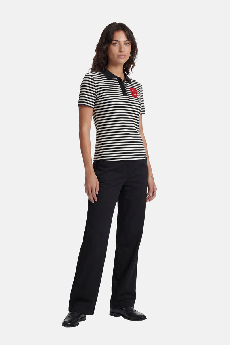 Vor einem hellgrauen Hintergrund steht eine Frau mit schulterlangem, gewelltem Haar und ausdruckslosem Gesicht. Sie trägt ein gestreiftes T-Shirt der Marke „Princess Goes Hollywood“ mit einer Snoopy-Stickerei, eine schwarze Hose und schwarze Schuhe. Ihre Arme liegen entspannt an den Seiten.