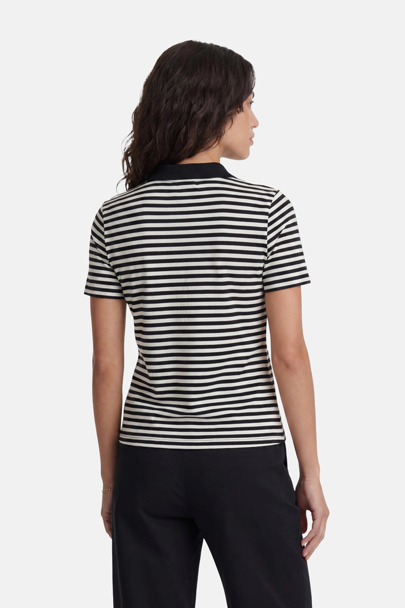 Eine Frau mit welligem, dunklem Haar steht mit dem Rücken zur Kamera und trägt ein schwarz-weiß gestreiftes T-Shirt aus der limitierten Auflage „Princess Goes Hollywood“ sowie eine schwarze Hose. Der schlichte, helle Hintergrund bringt ihr exklusives Outfit besonders gut zur Geltung.