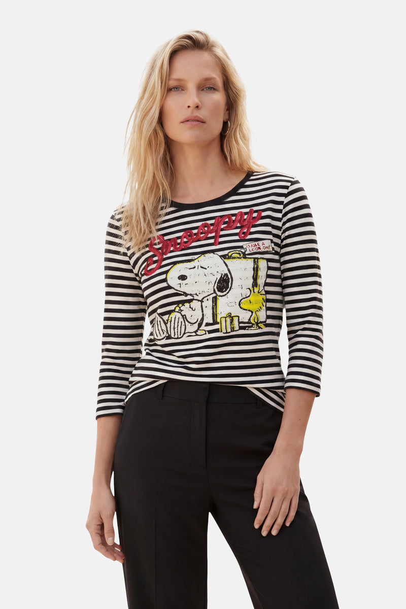 Eine Frau mit welligem blondem Haar trägt ein „Princess Goes Hollywood Limited Edition Snoopy“-Shirt – ein schwarz-weiß gestreiftes Oberteil mit 3/4-Ärmeln, auf dem Snoopy und Woodstock abgebildet sind, sowie den roten Schriftzug „Snoopy“ – und eine schwarze Hose. Sie steht selbstbewusst vor einem schlichten Hintergrund.