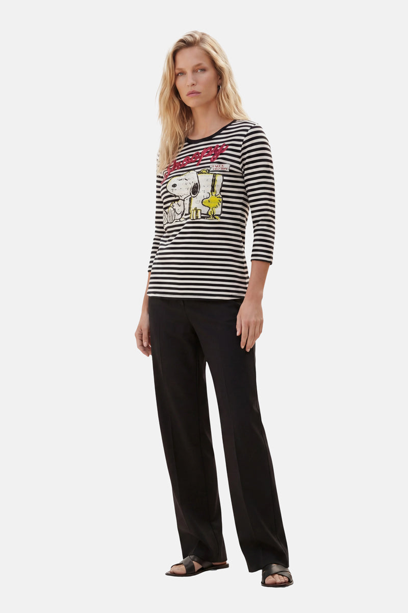 Eine Frau mit langen blonden Haaren trägt ein Snoopy-Shirt aus der „Princess Goes Hollywood“-Limited-Edition – aus schwarz-weiß gestreifter Baumwolle mit Snoopy und Woodstock –, dazu schwarze Hosen und Sandalen. Sie steht mit neutralem Gesichtsausdruck vor einem hellgrauen Hintergrund und blickt geradeaus.
