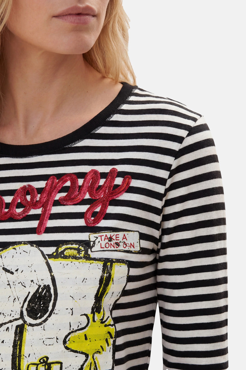 Eine Frau mit heller Haut und welligem blondem Haar trägt ein „Princess Goes Hollywood Limited Edition Snoopy“-Shirt – ein schwarz-weiß gestreiftes Langarm-Oberteil aus Baumwolle mit dem roten Schriftzug „Happy“, Snoopy- und Woodstock-Motiven sowie einem „TAKE A LONDON“-Aufnäher. Weißer Hintergrund.