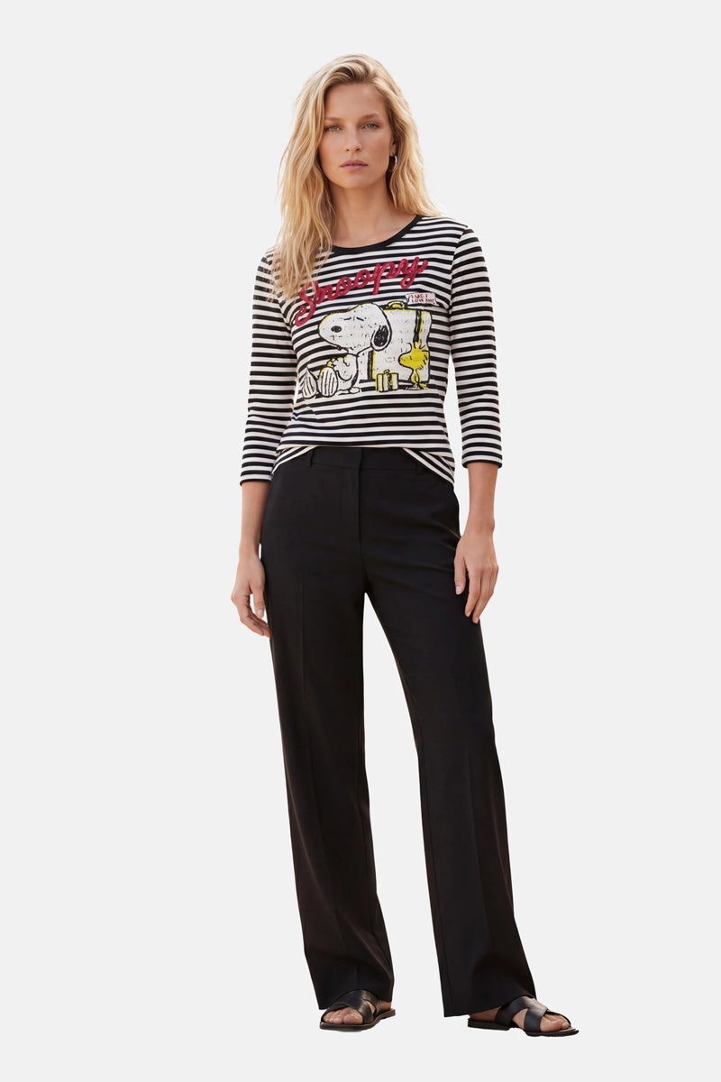 Eine Frau mit langen blonden Haaren und ausdruckslosem Gesicht präsentiert vor einem schlichten Hintergrund ein schwarz-weiß gestreiftes Snoopy-Shirt aus der Limited Edition von „Princess Goes Hollywood“, kombiniert mit einer schwarzen Hose mit weitem Bein und Sandalen.