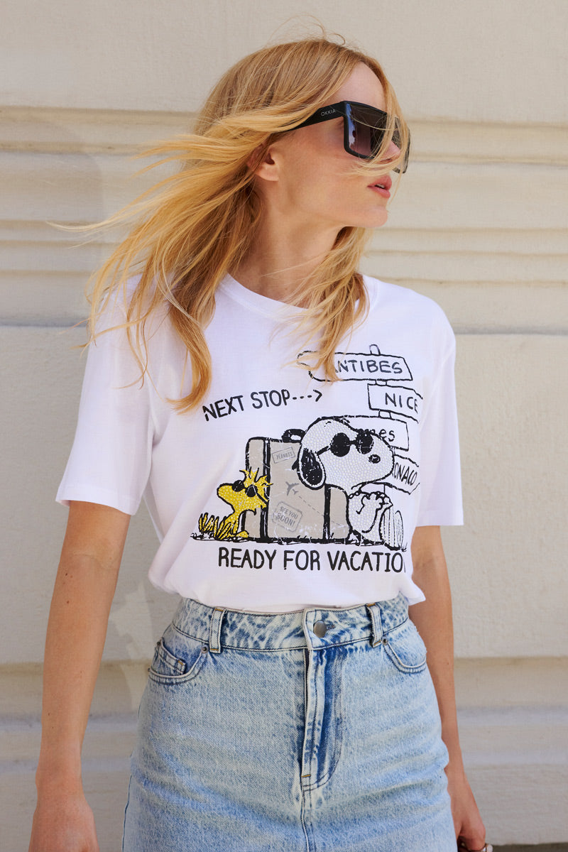 Eine Frau mit langen blonden Haaren trägt eine große schwarze Sonnenbrille, ein Snoopy-T-Shirt aus Baumwolle der Limited Edition „Princess Goes Hollywood“ mit der Aufschrift „Next stop... Ready for vacation“ sowie einen hellblauen Jeansrock mit hoher Taille und steht vor einer cremefarbenen, strukturierten Wand.