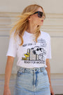 Eine Frau mit langen blonden Haaren trägt eine große schwarze Sonnenbrille, ein Snoopy-T-Shirt aus Baumwolle der Limited Edition „Princess Goes Hollywood“ mit der Aufschrift „Next stop... Ready for vacation“ sowie einen hellblauen Jeansrock mit hoher Taille und steht vor einer cremefarbenen, strukturierten Wand.