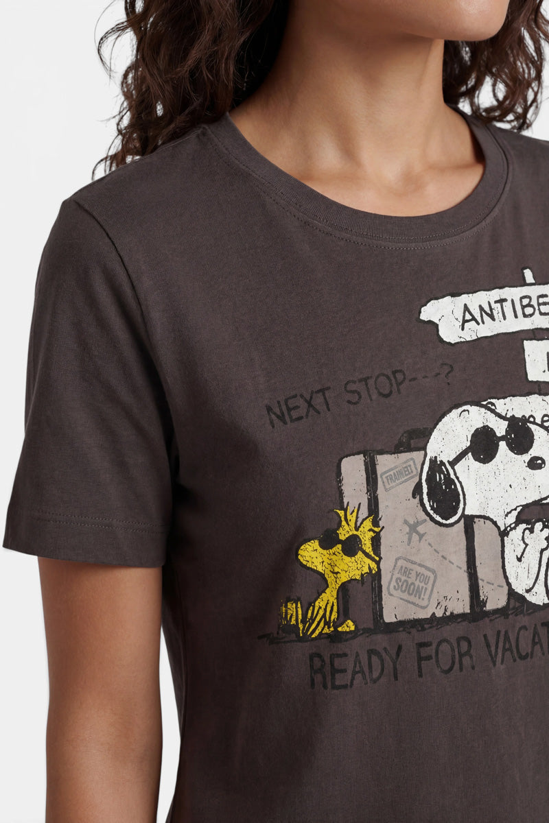 Eine Frau trägt ein Snoopy-T-Shirt aus Baumwolle der Limited Edition „Princess Goes Hollywood“, auf dem Snoopy mit Sonnenbrille, Woodstock und Vintage-Koffern abgebildet sind. Text: „Nächster Halt…? Antibes. Bereit für den Urlaub.“ Zu sehen sind nur ihr unterer Gesichtsbereich und ihr Oberkörper.