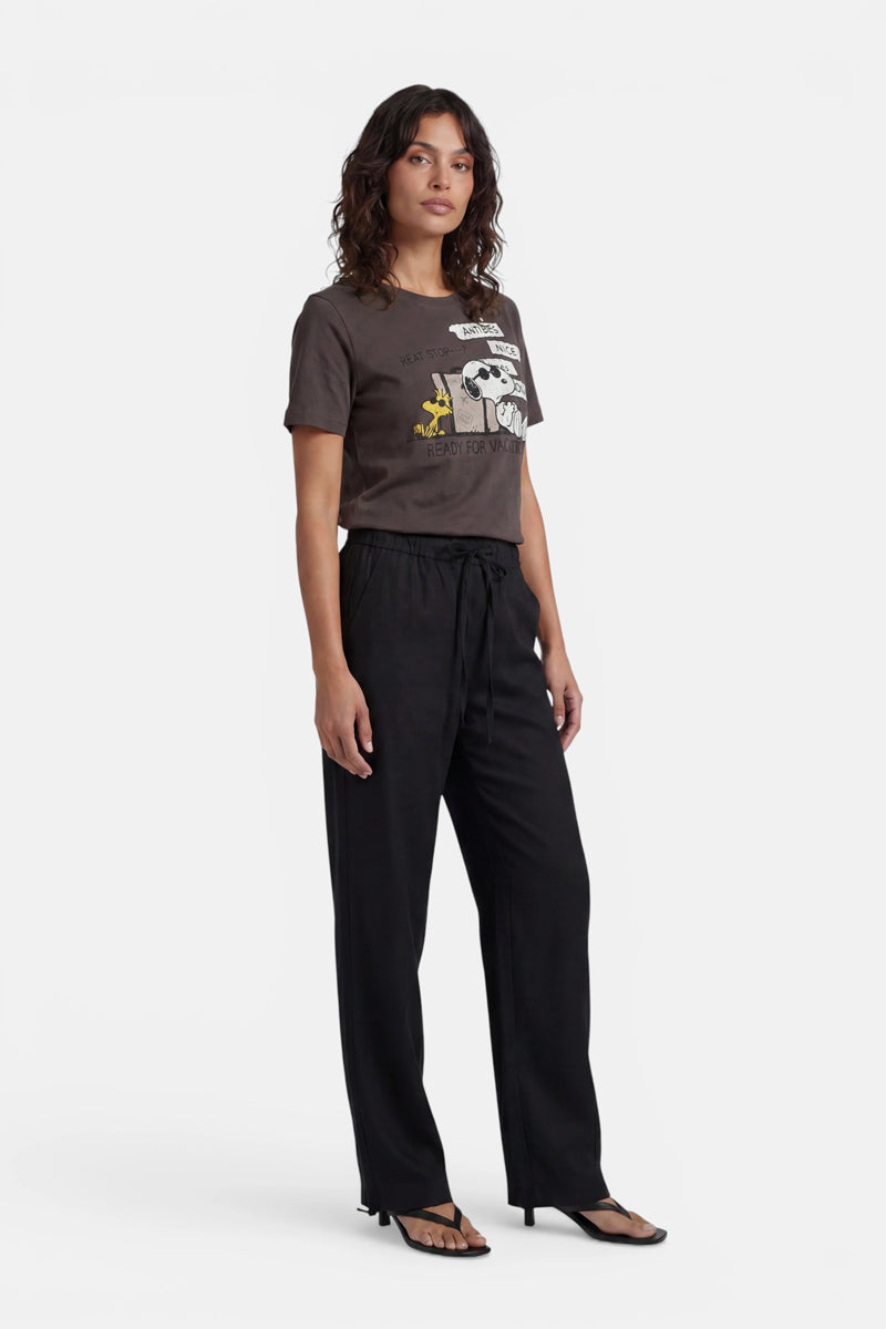 Eine Frau mit welligem, dunklem Haar steht vor einem weißen Hintergrund. Sie trägt das „Princess Goes Hollywood“-Snoopy-T-Shirt in limitierter Auflage mit gelben Akzenten, eine lockere schwarze Hose mit Kordelzug und schwarze Sandalen. Ihr Gesichtsausdruck ist neutral und ihre Körperhaltung entspannt.